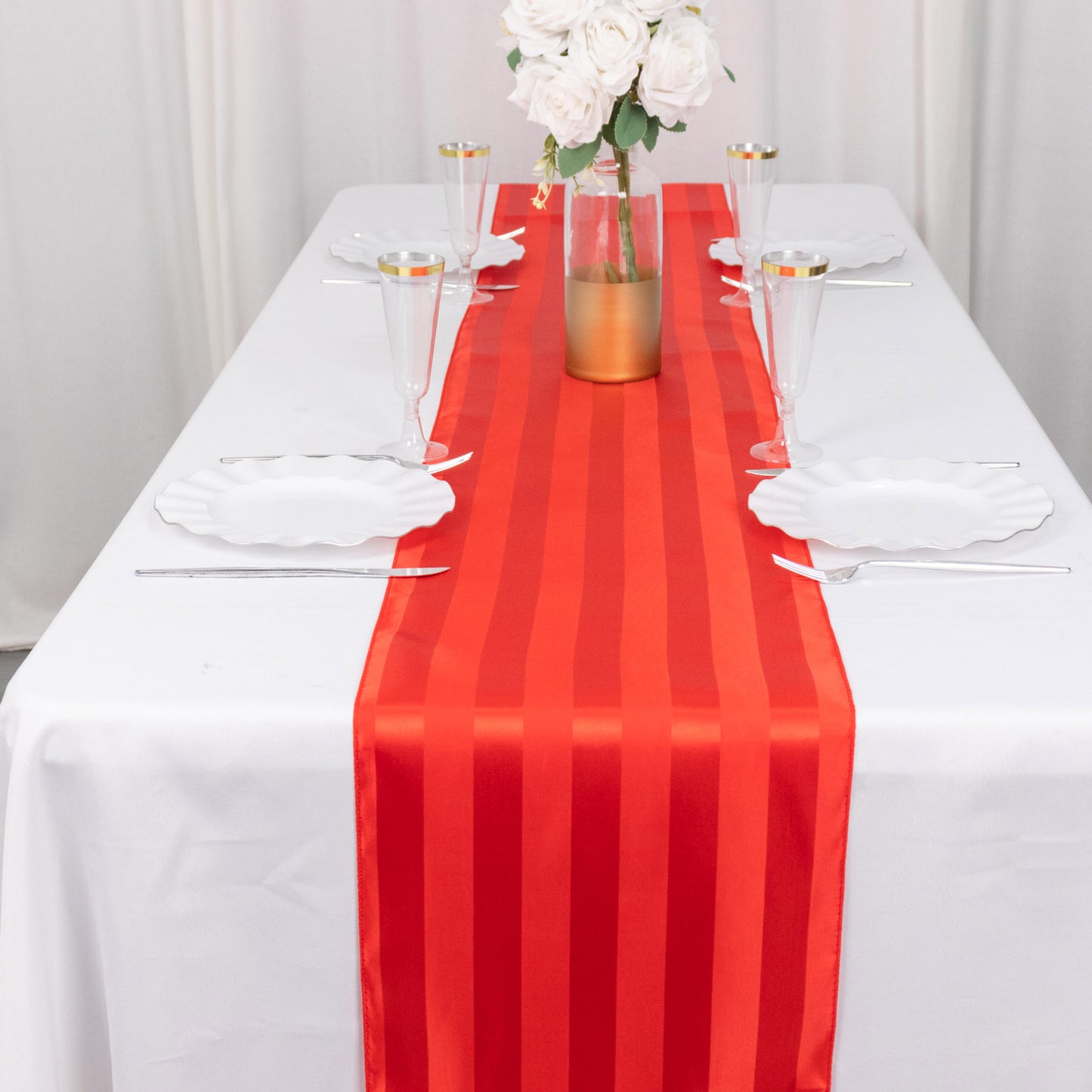 Red satin stripe table runner on a white table set with flowers and dishes.