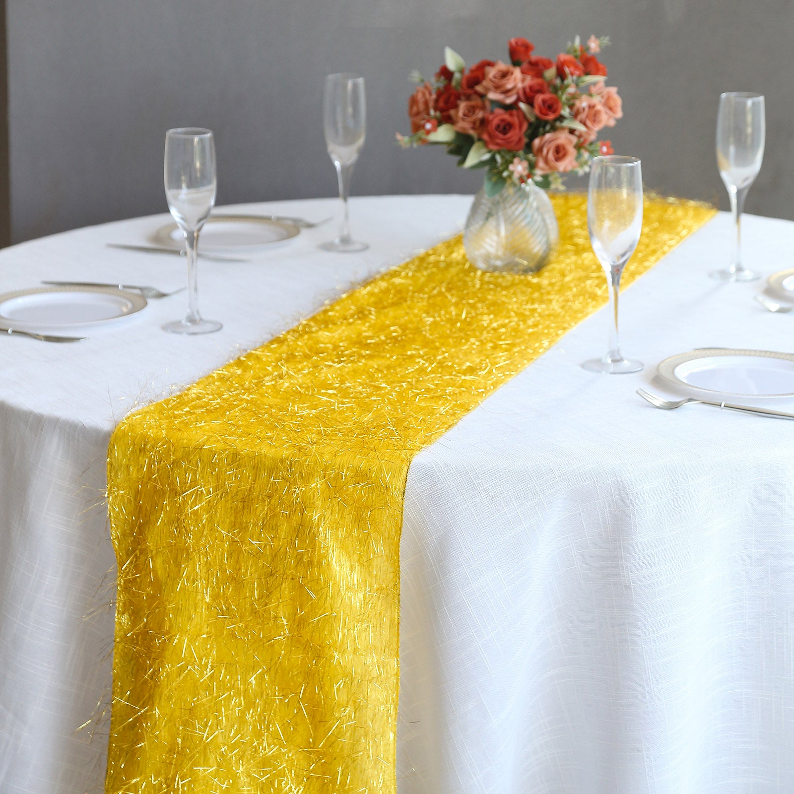 Gold metallic fringe table runner on a white table, surrounded by glassware and flowers.