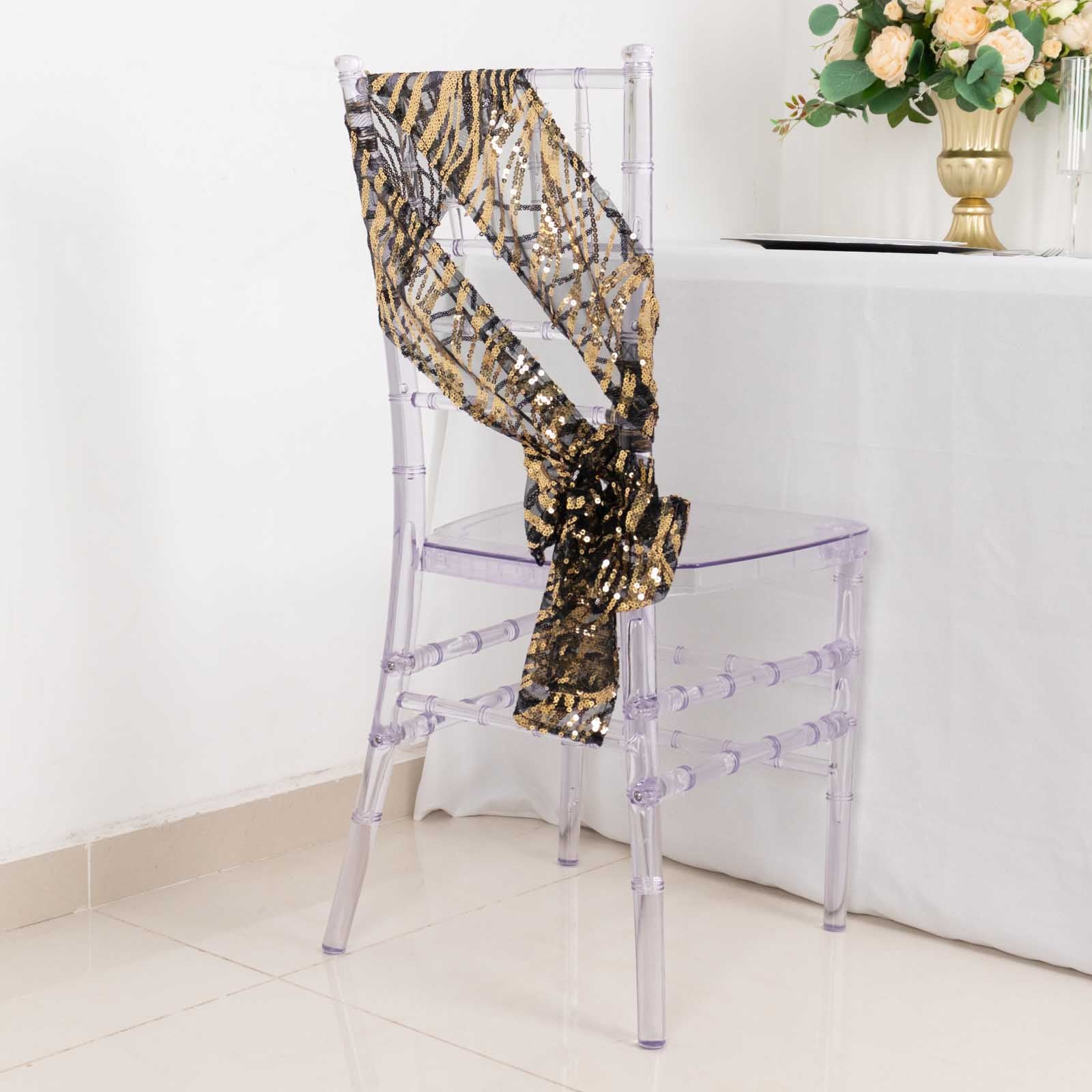 Black and gold embroidered sequin mesh chair sash on a clear chair.