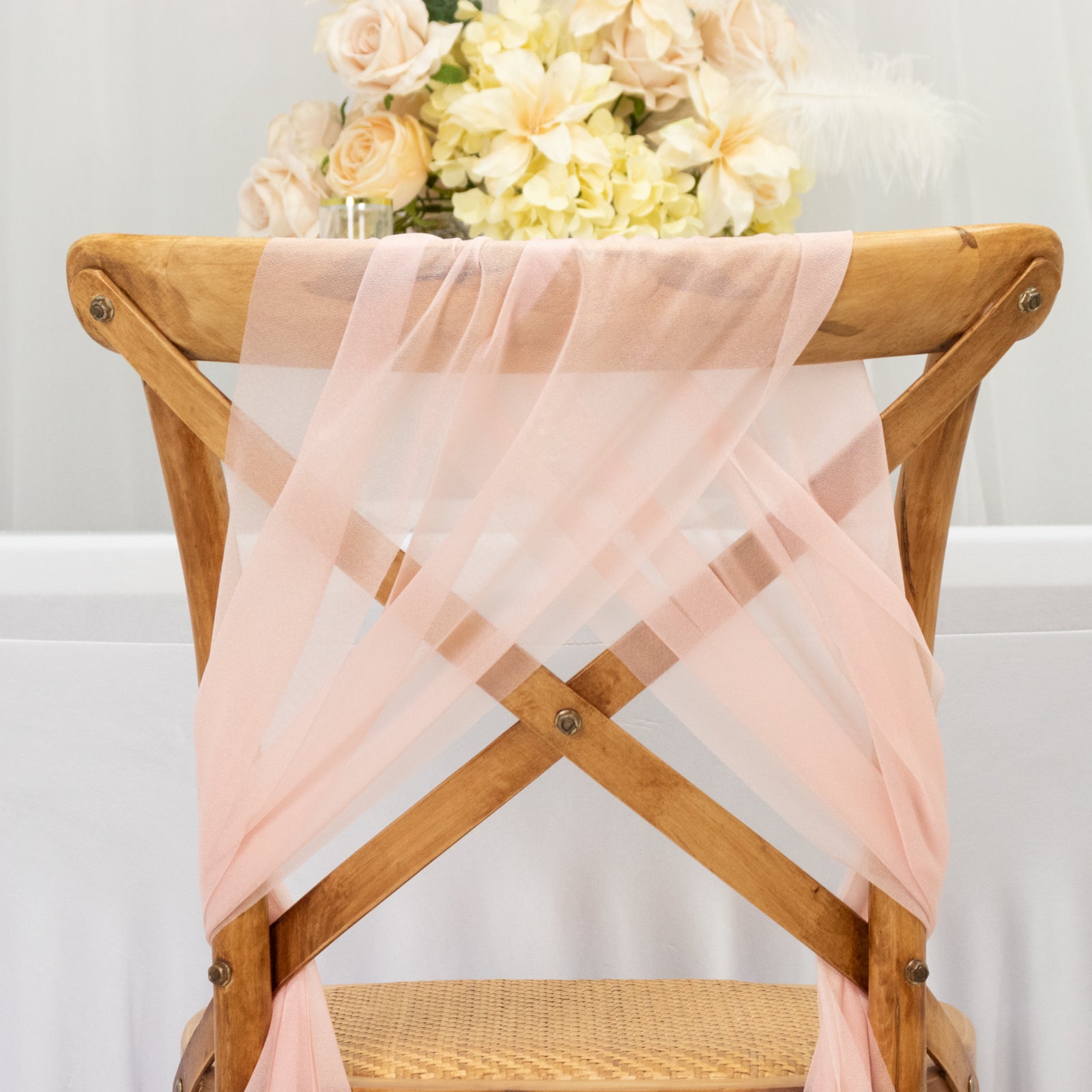 Blush sheer chair drape decor on a wooden chair with floral arrangement backdrop.