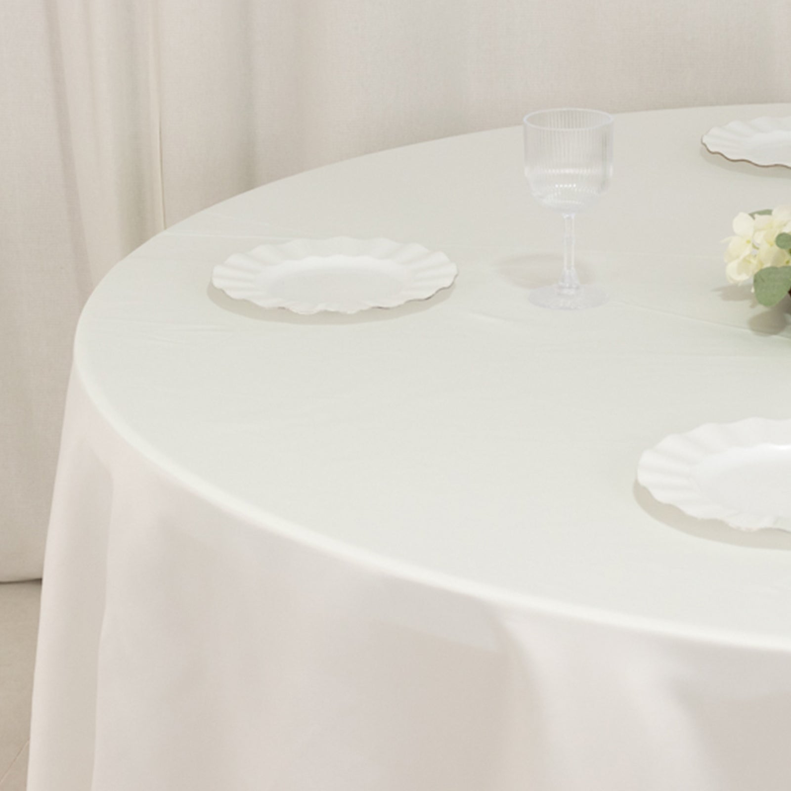 Ivory round satin tablecloth on a dining table with plates and a glass.