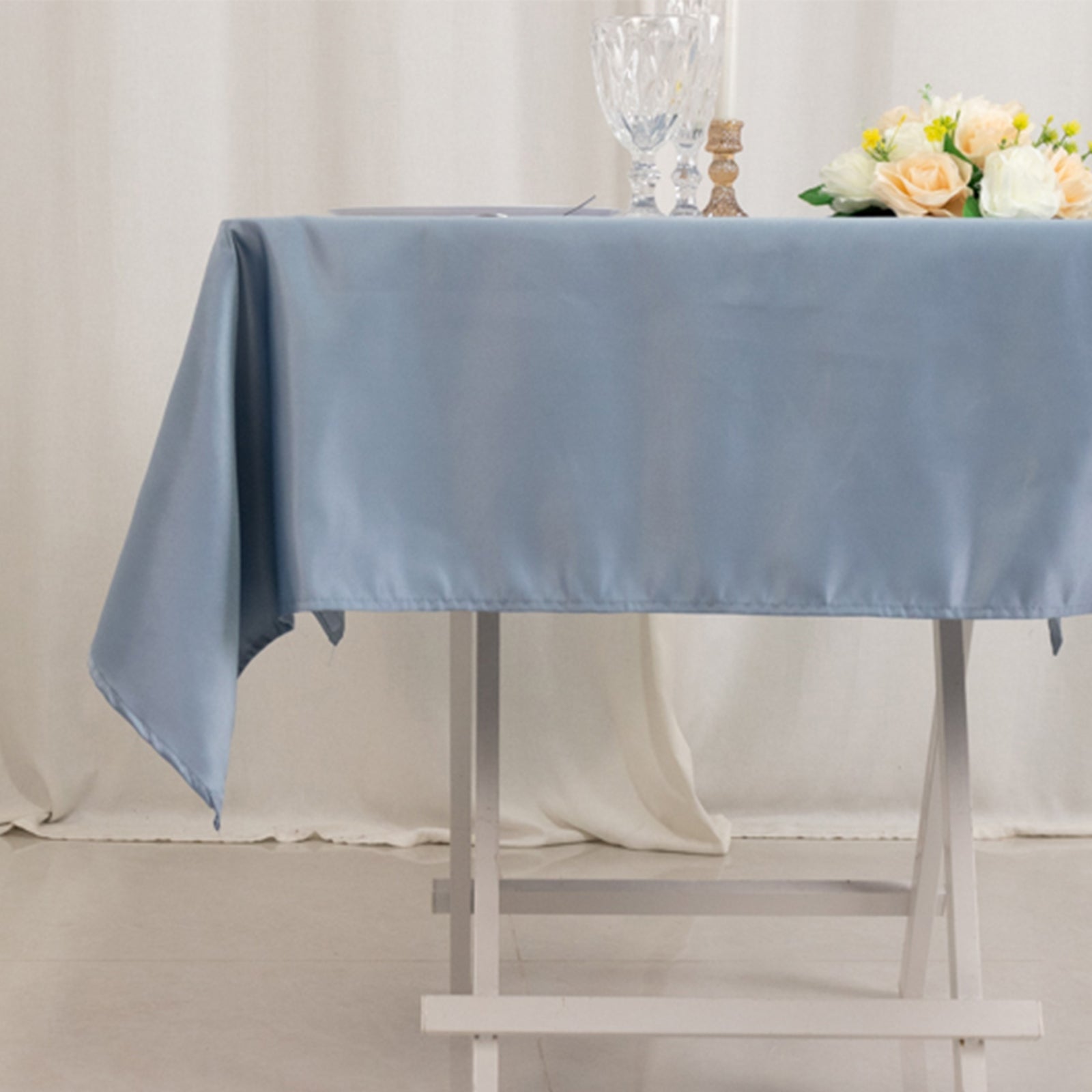 Dusty blue satin square tablecloth on a table with flowers and glassware.