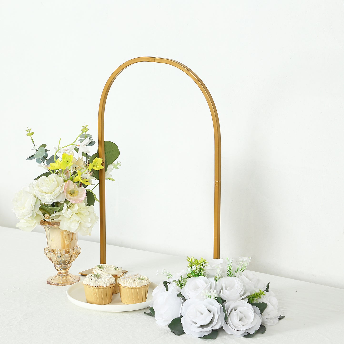 Gold Chiara arch wedding cake display stand with floral arrangements and cupcakes.
