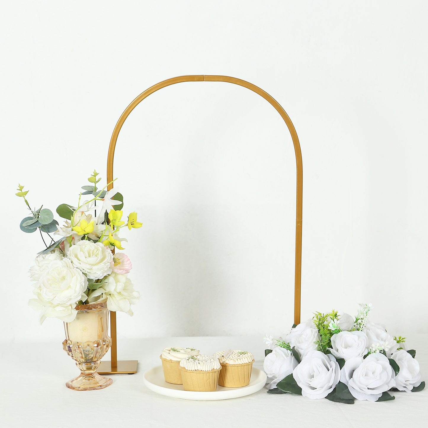 Gold metal arch wedding cake display stand with floral centerpiece and cupcakes.