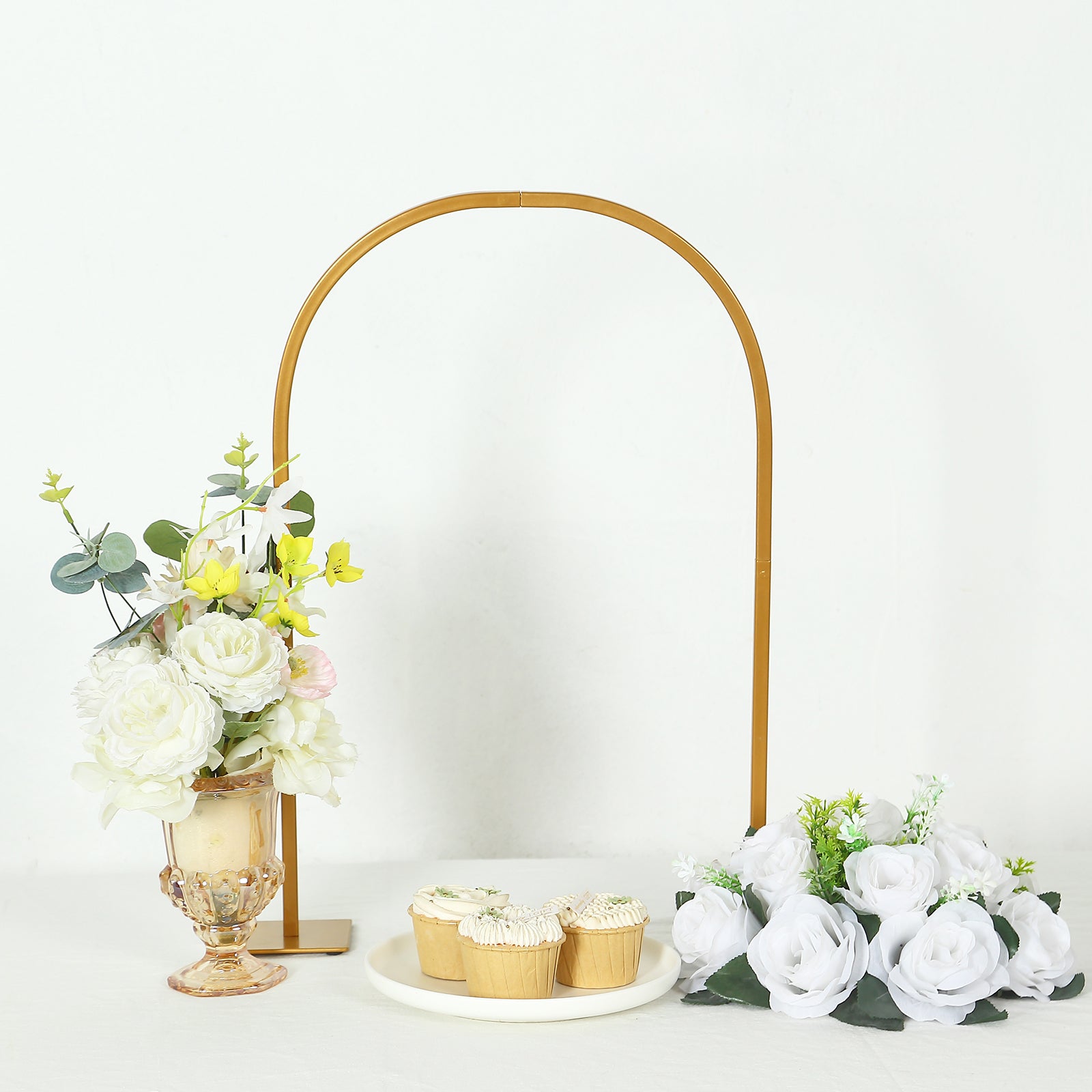 Gold metal arch wedding cake display stand with floral centerpiece and cupcakes.