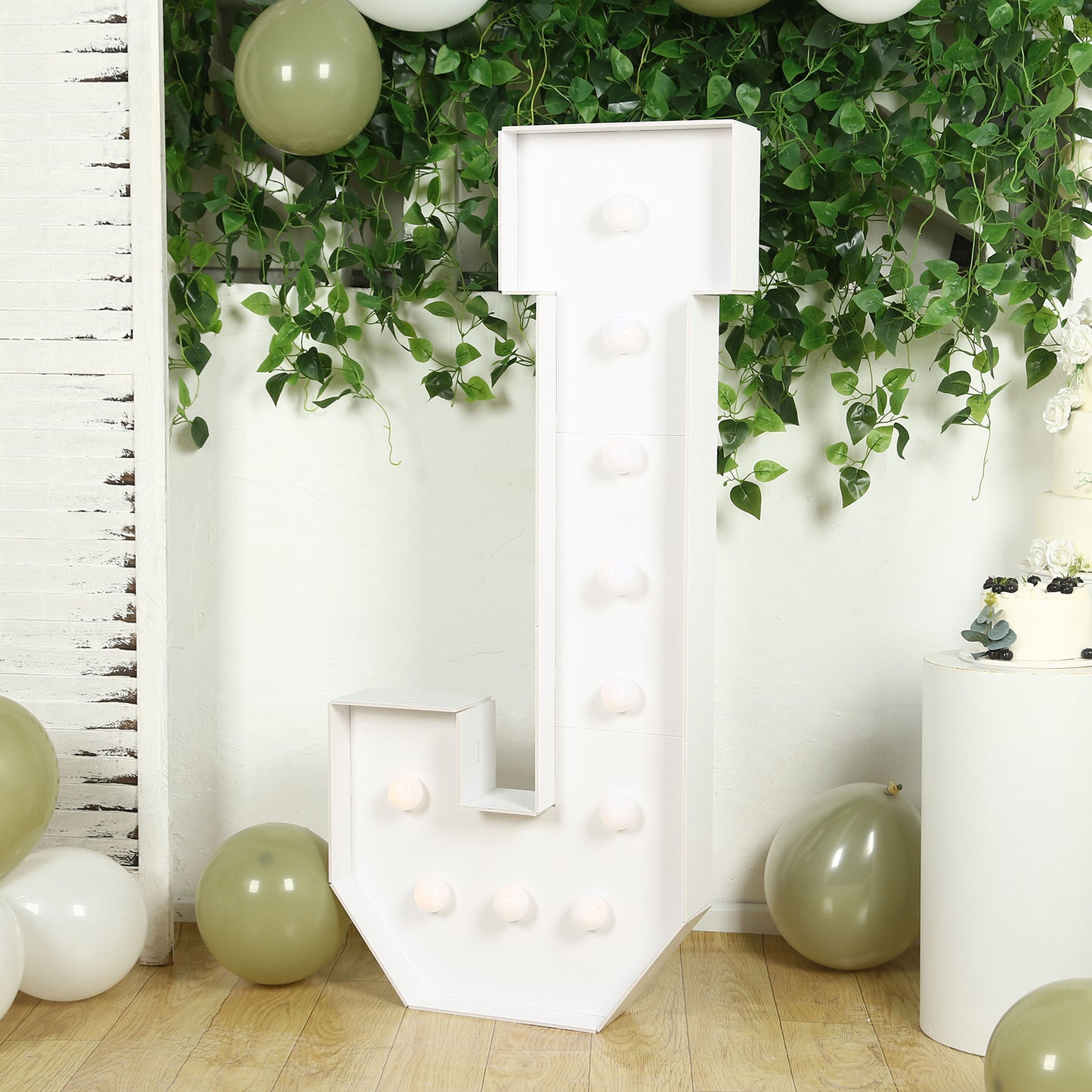 White LED marquee letter "J" with warm white lights, surrounded by greenery and balloons.