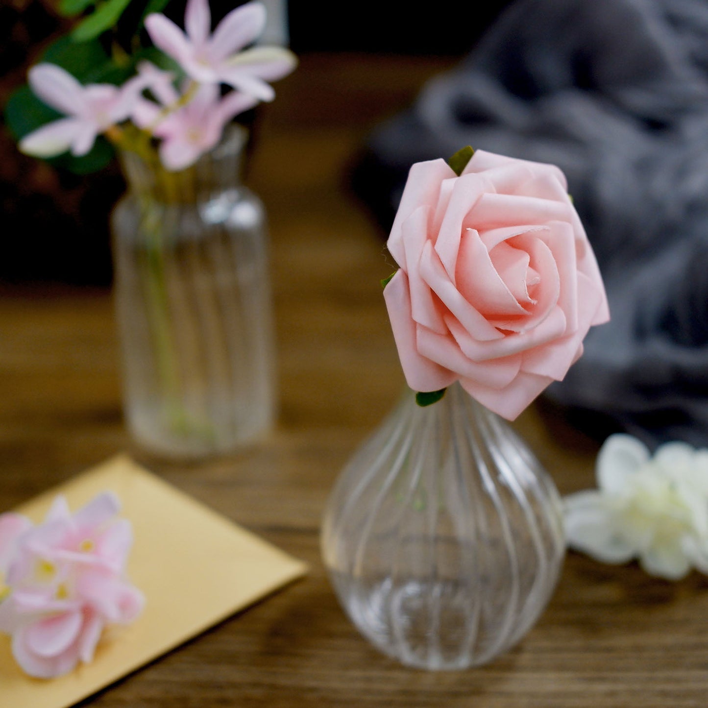 Pink artificial rose in a glass vase with assorted flowers on a wooden table.