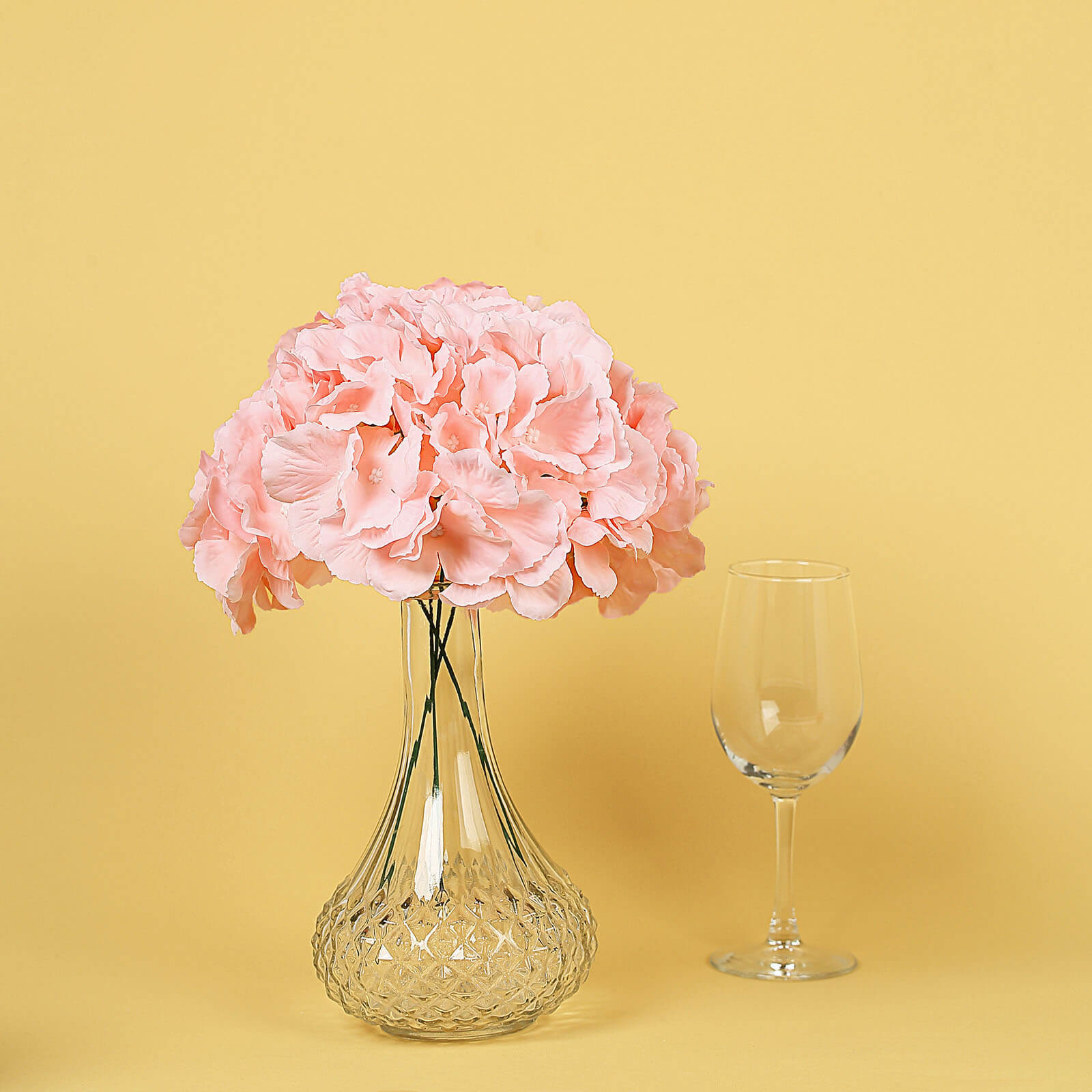 Blush artificial satin hydrangeas in a glass vase on a yellow background.