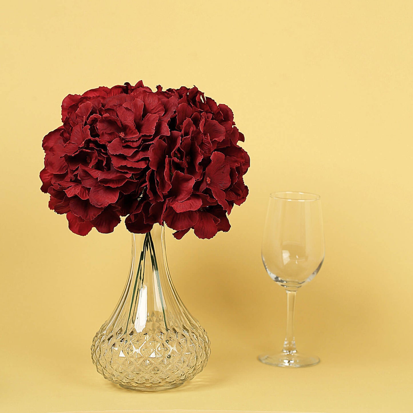 Burgundy artificial satin hydrangeas in a glass vase on a yellow background.