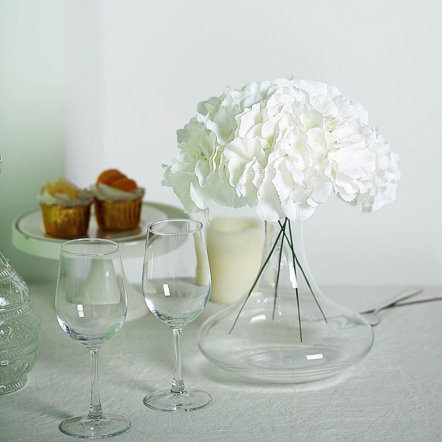 Cream artificial satin hydrangeas in a clear vase with two wine glasses.