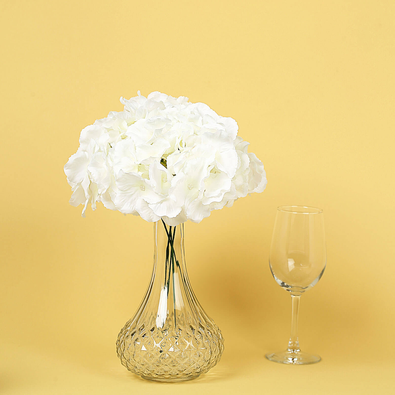 Cream artificial satin hydrangeas in a glass vase on a yellow background.