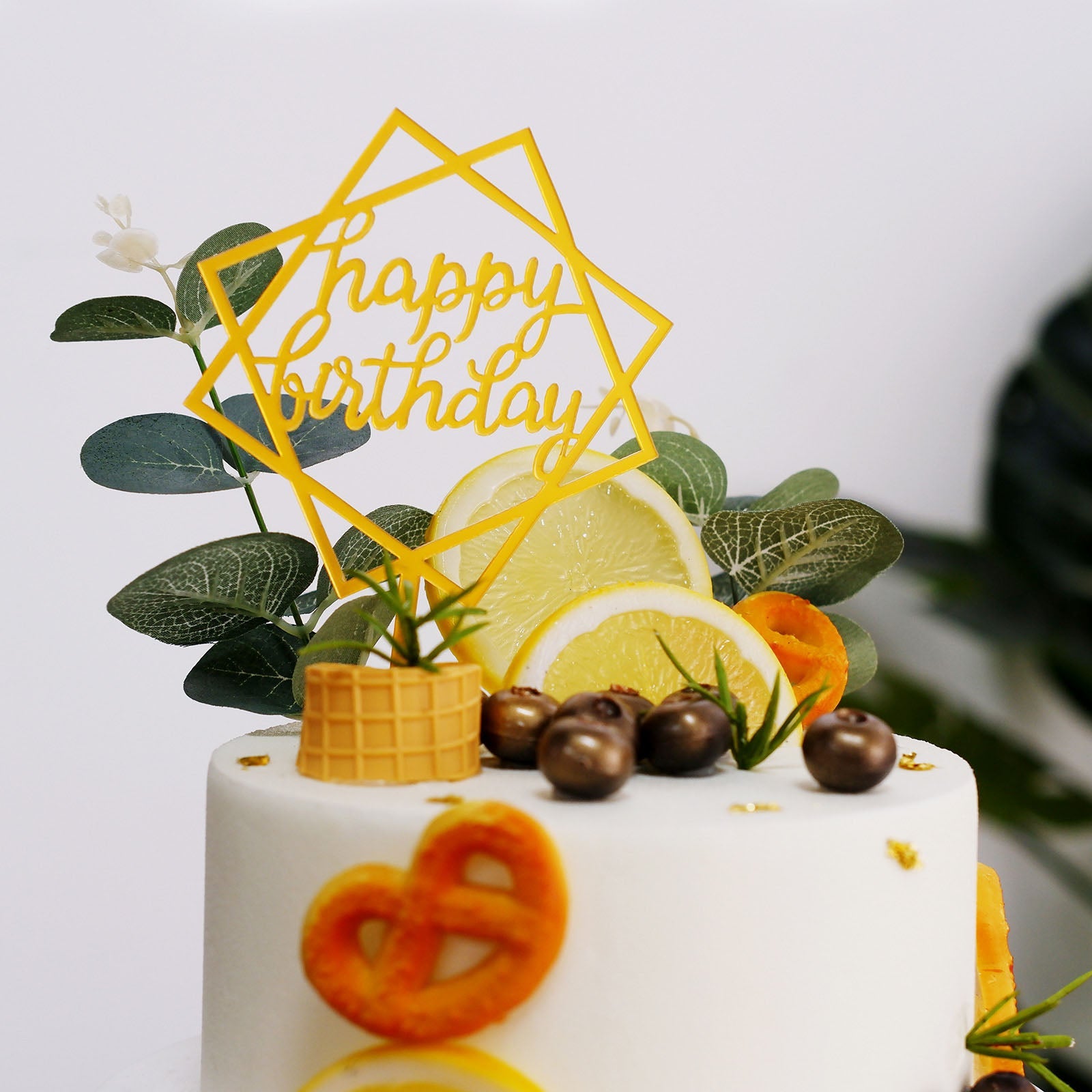Gold acrylic birthday cake topper on a decorated white cake with fruit and greenery.