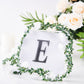 Black decorative rhinestone letter sticker "E" surrounded by greenery and flowers.