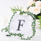 Black decorative rhinestone letter "F" surrounded by greenery and white flowers.