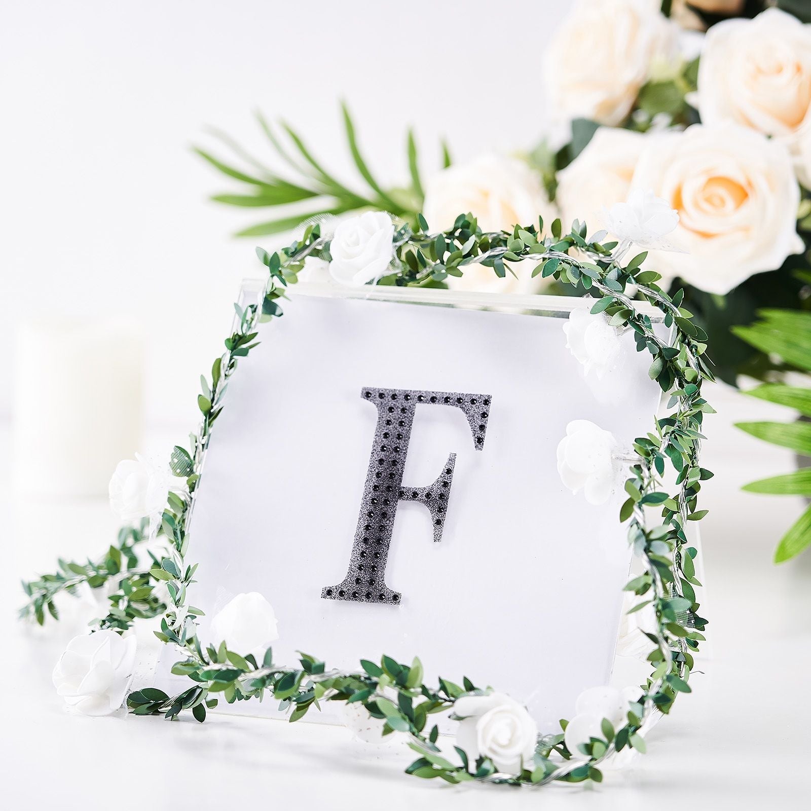 Black decorative rhinestone letter "F" surrounded by greenery and white flowers.