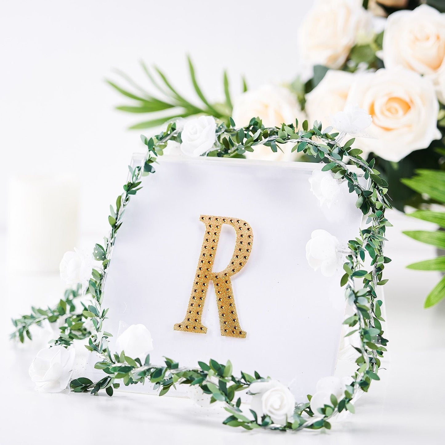 Gold decorative rhinestone letter sticker "R" surrounded by greenery and flowers.