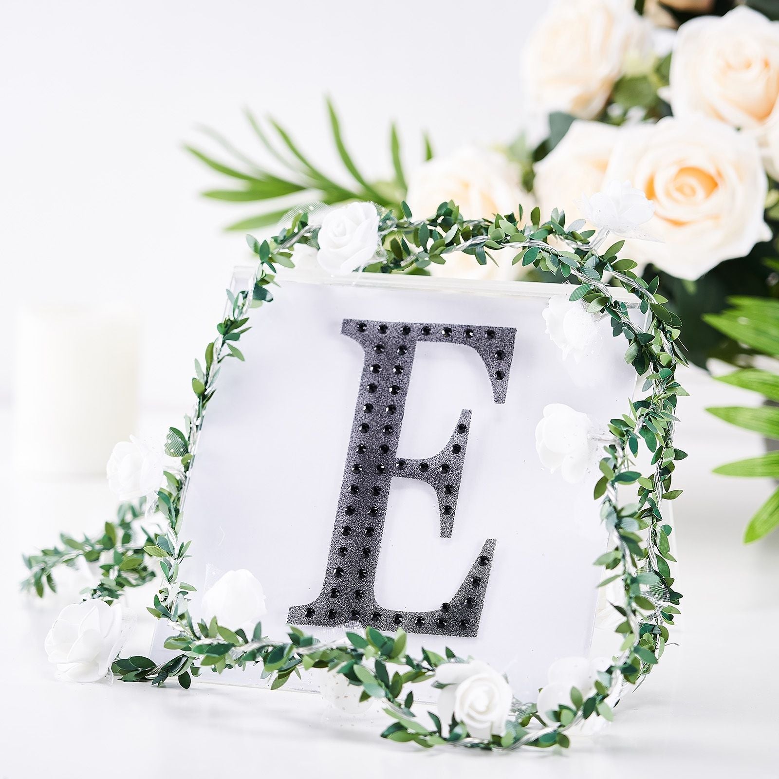 Black rhinestone letter "E" surrounded by greenery and flowers.