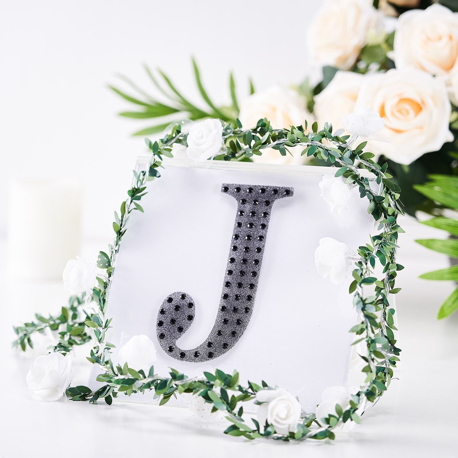Black rhinestone letter "J" surrounded by greenery and flowers.