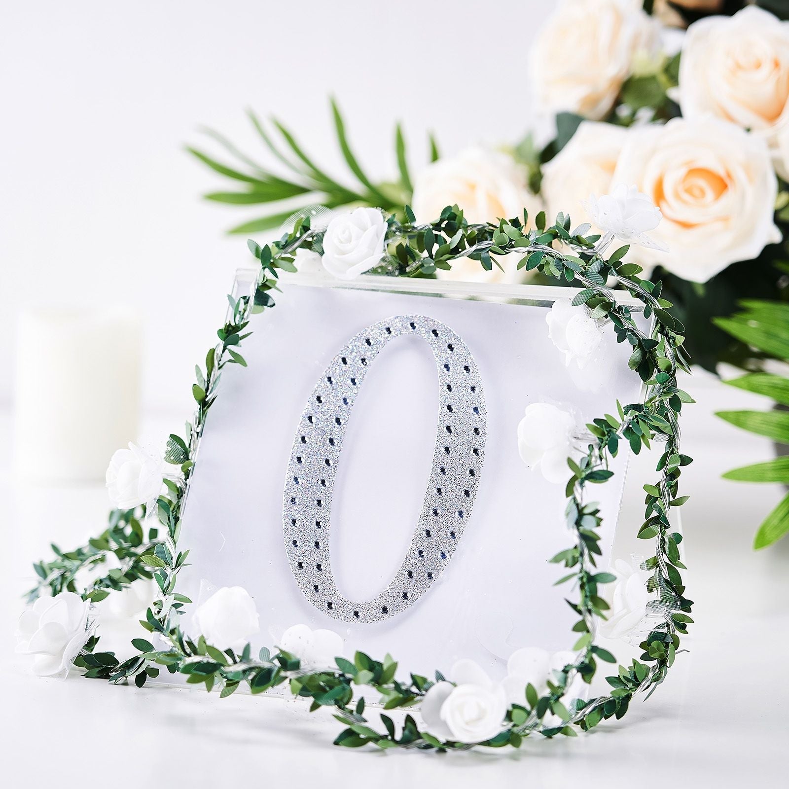 Silver decorative rhinestone letter "O" surrounded by greenery and white flowers.