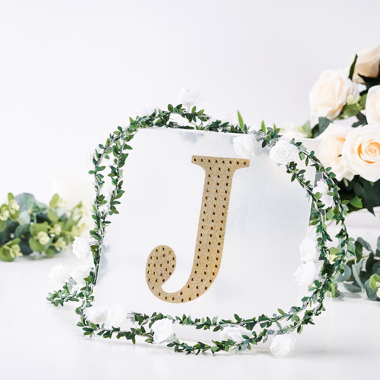 Gold decorative rhinestone letter "J" surrounded by greenery and flowers.