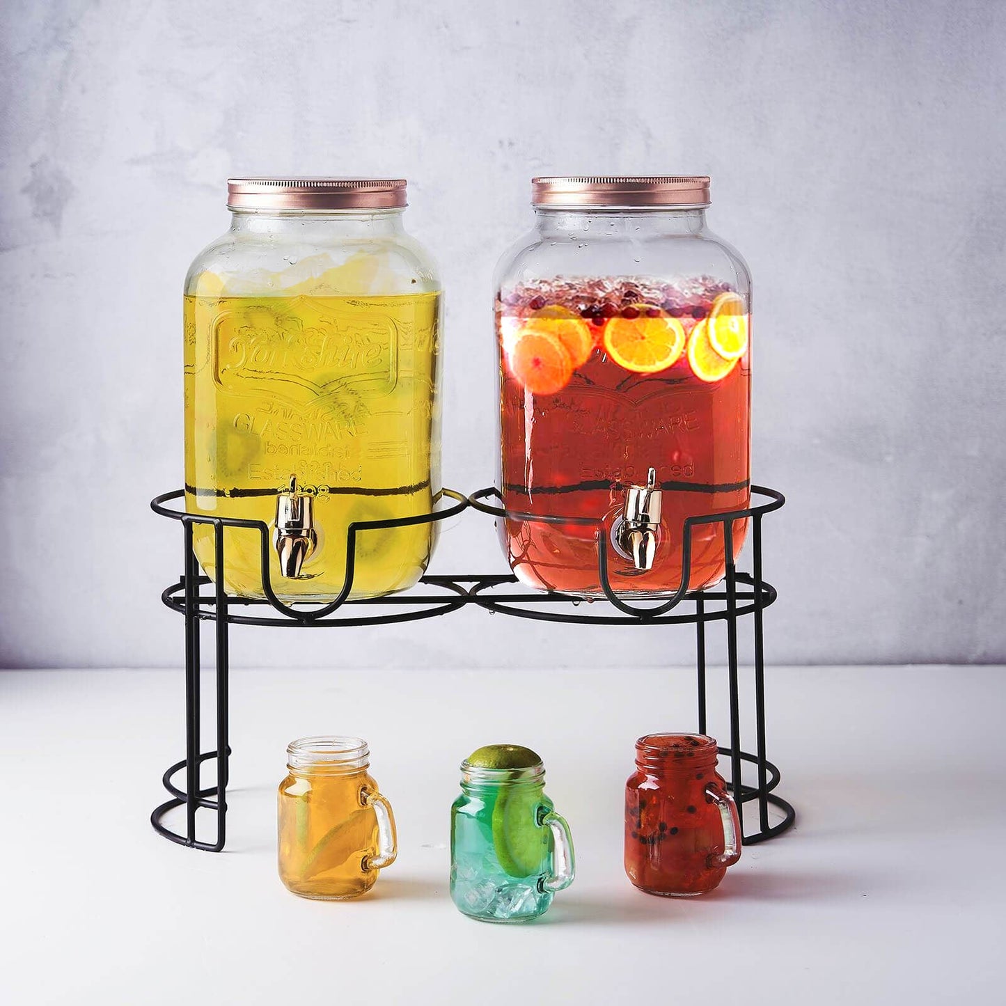 Clear dual gallon glass jars with rose gold lids on a black stand.