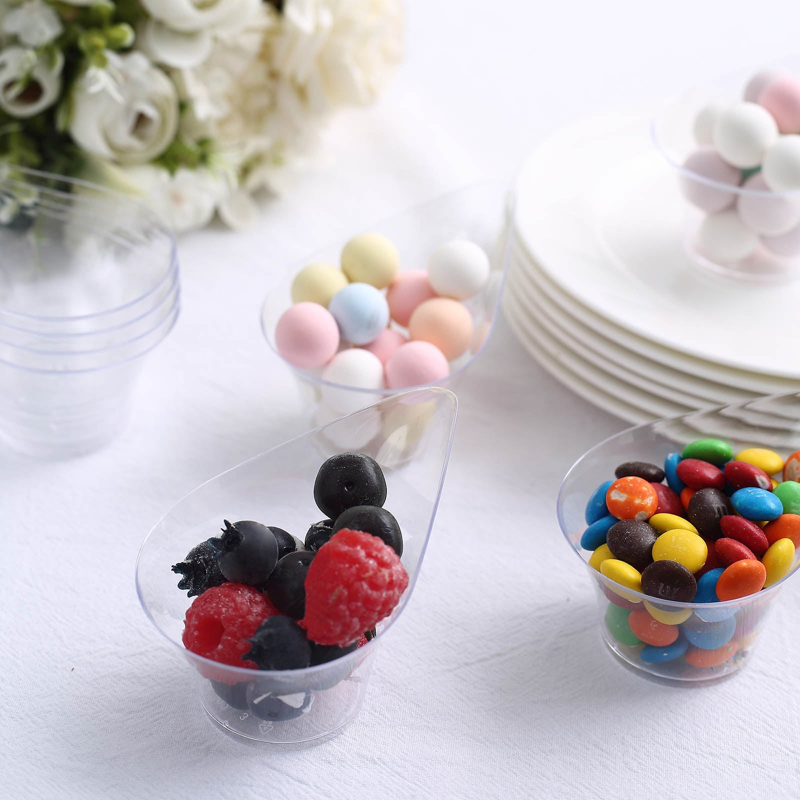 Clear mini disposable teardrop dessert cups with colorful snacks inside.