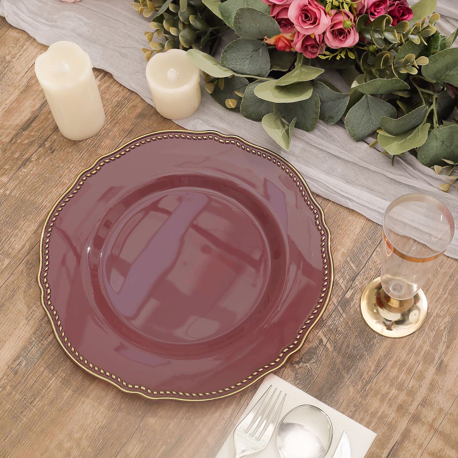 Elegant burgundy plate with gold trim, candles, and floral arrangement on a rustic table.