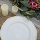 Elegant white plate with gold trim beside candles and floral arrangement on a wooden table.