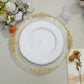 Elegant white plate with gold trim on decorative charger, surrounded by floral decor.