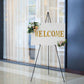 65" black metal easel stand displaying a welcome sign with floral accents.