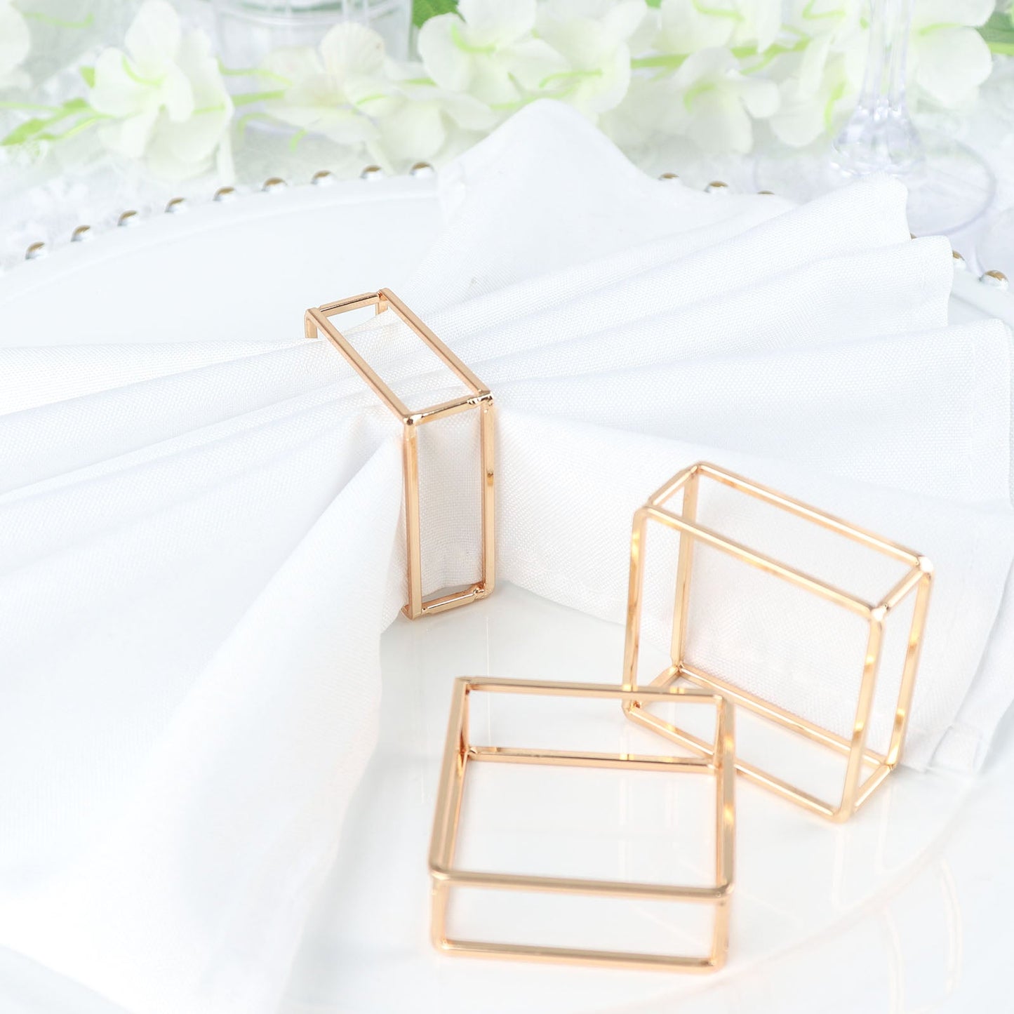 Gold metal hollow square napkin rings on a white table setting.