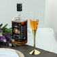 Clear champagne flute with gold base beside a bottle of whiskey and flowers.