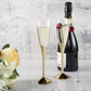 Clear champagne flutes with gold bases, featuring cherries, next to a champagne bottle.