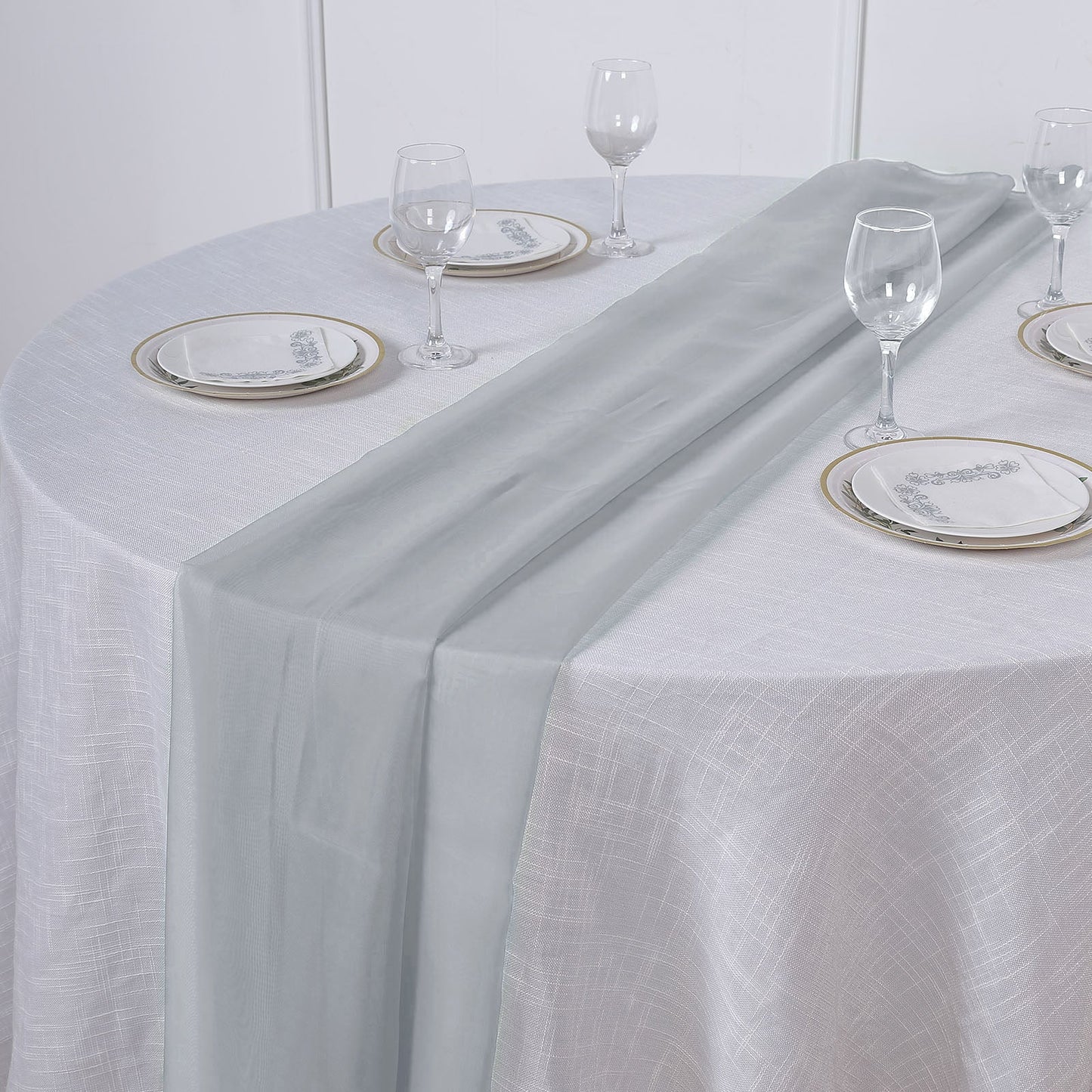Silver chiffon table runner on a round table set with glassware and plates.