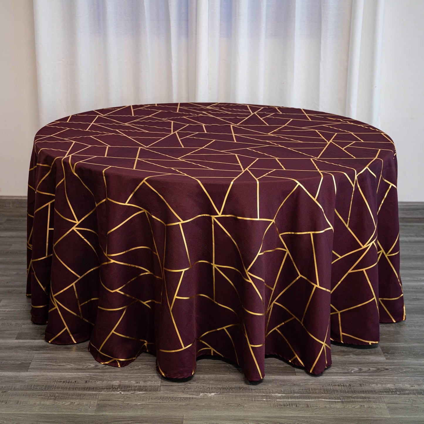 Burgundy round tablecloth with gold geometric pattern on a wooden floor.