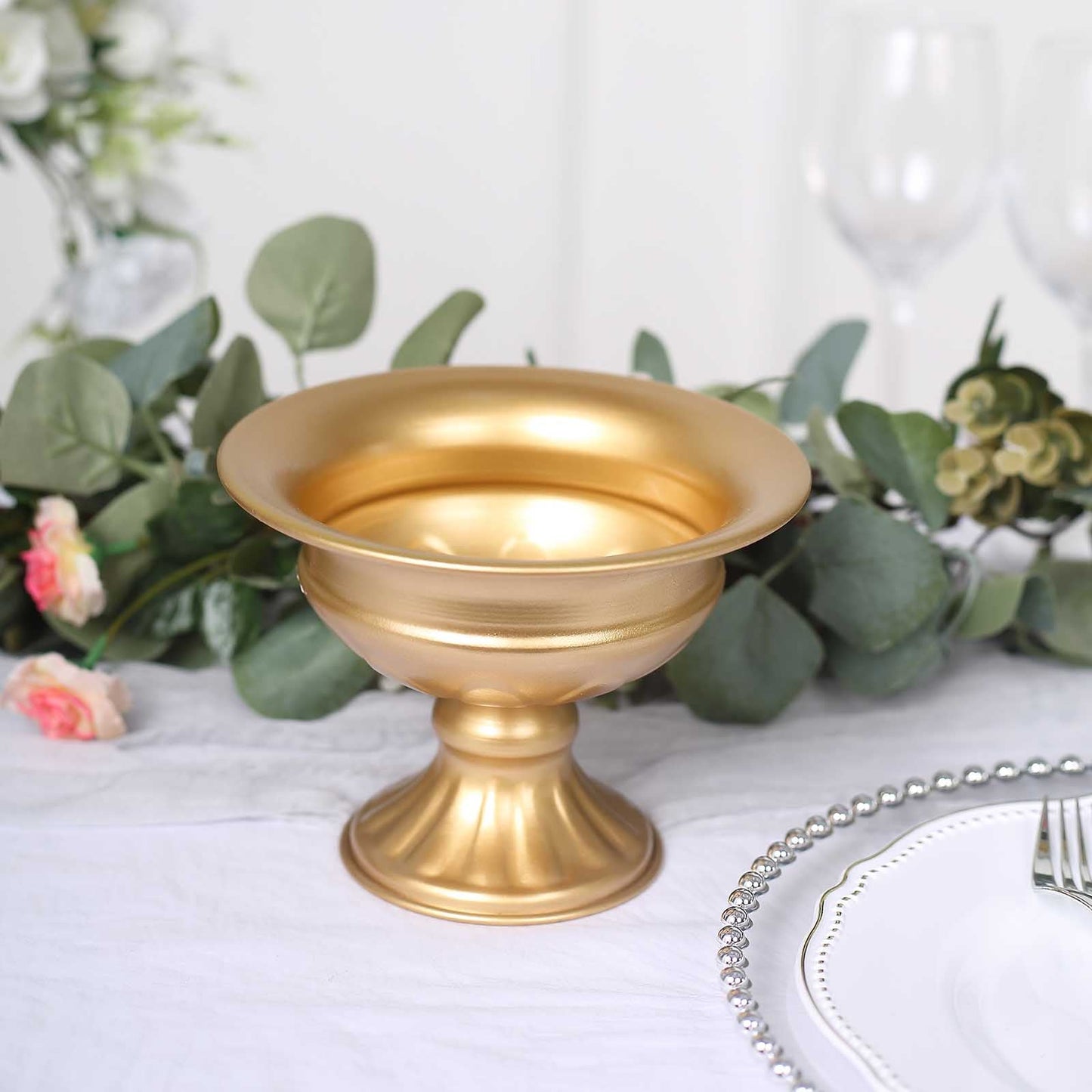Gold metal flower pedestal vase on a table with greenery and floral accents.