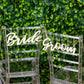Natural wood bride and groom chair signs on clear chairs against greenery backdrop.