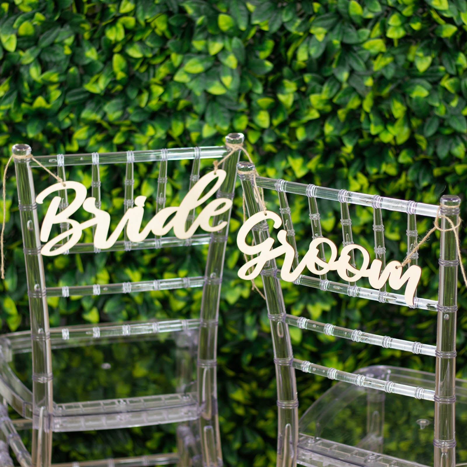 Natural wood bride and groom chair signs on clear chairs against greenery backdrop.